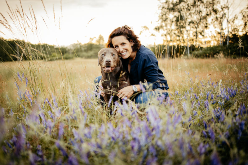 Eine Frau und ein Hund sitzen in einer Blumenwiese.